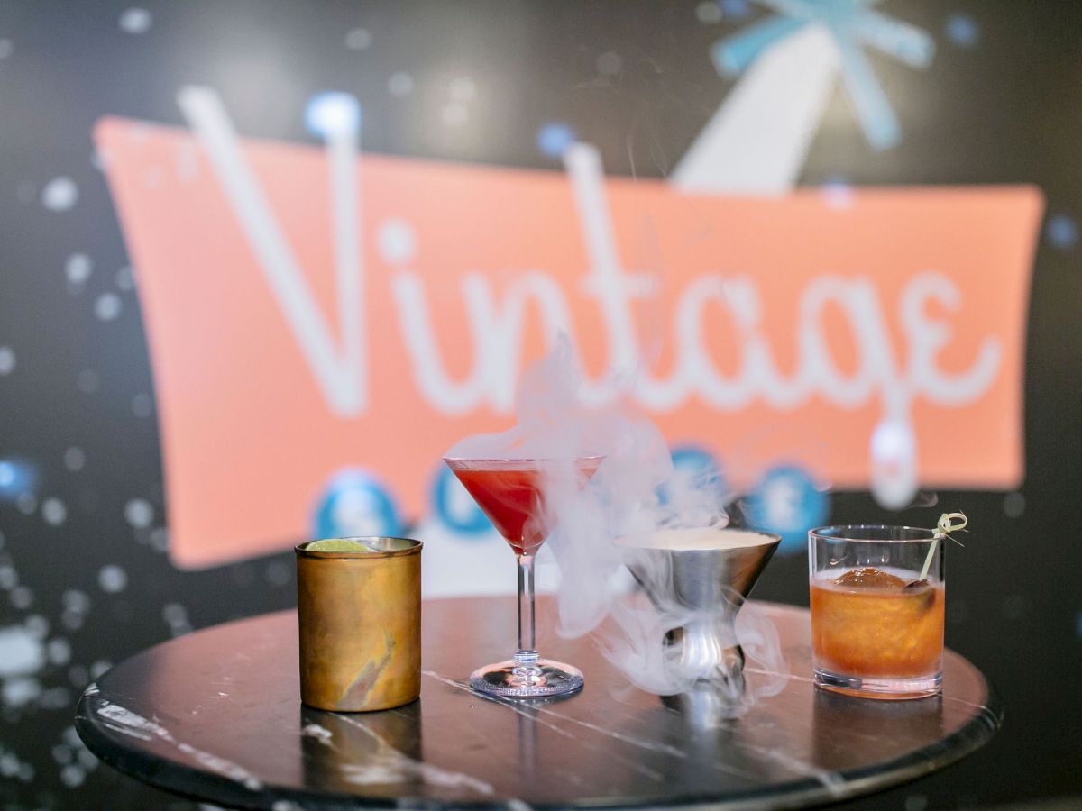 Four cocktails on a table with a blurred "Vintage" sign in the background, featuring playful smoke effects.