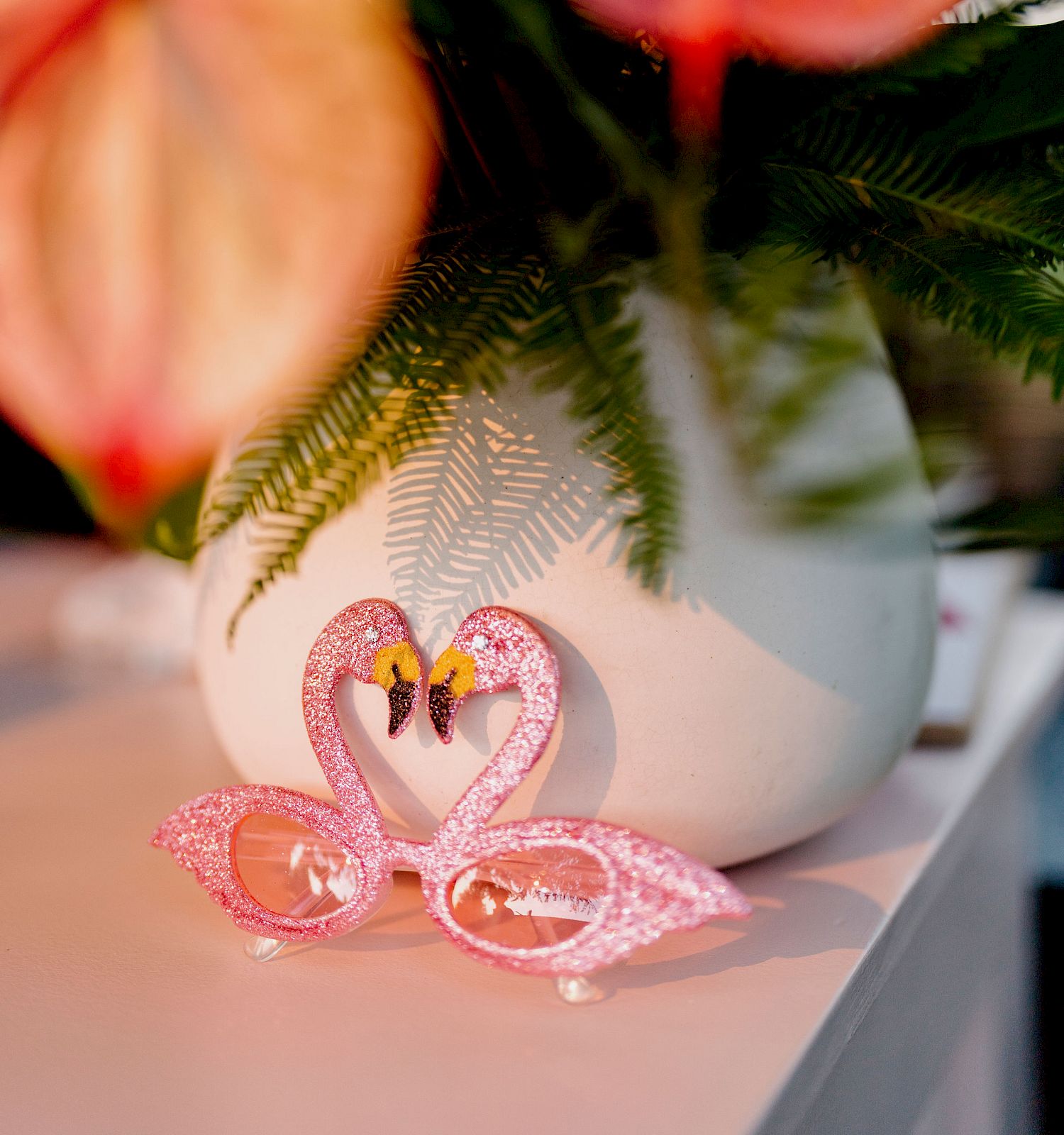 A pair of pink, flamingo-shaped sunglasses is placed on a table in front of a white vase with anthurium flowers and green leaves, creating a colorful display.