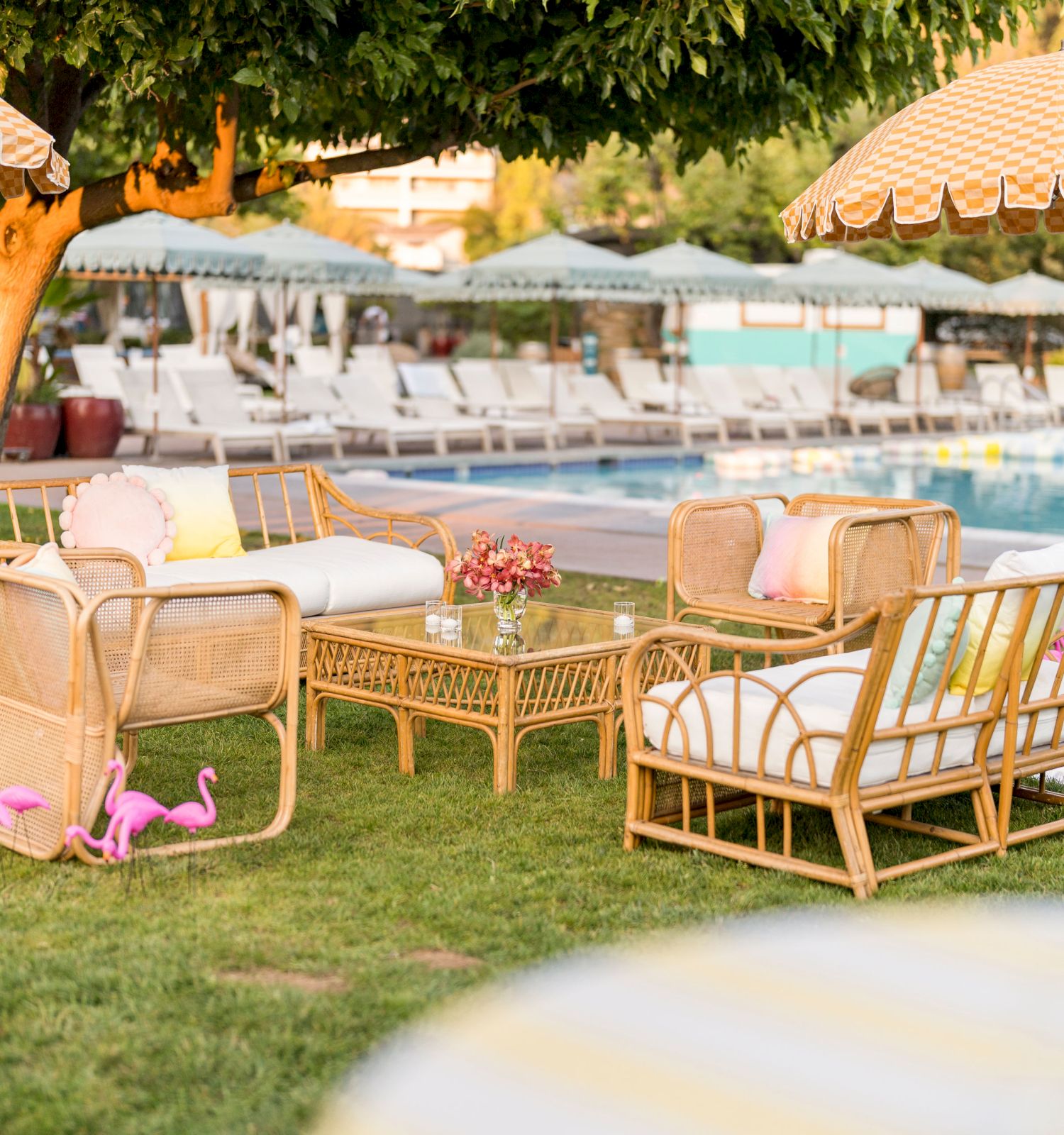 Outdoor seating area with wicker furniture, cushions, and a table set on grass near a pool with umbrellas and lounge chairs in the background.