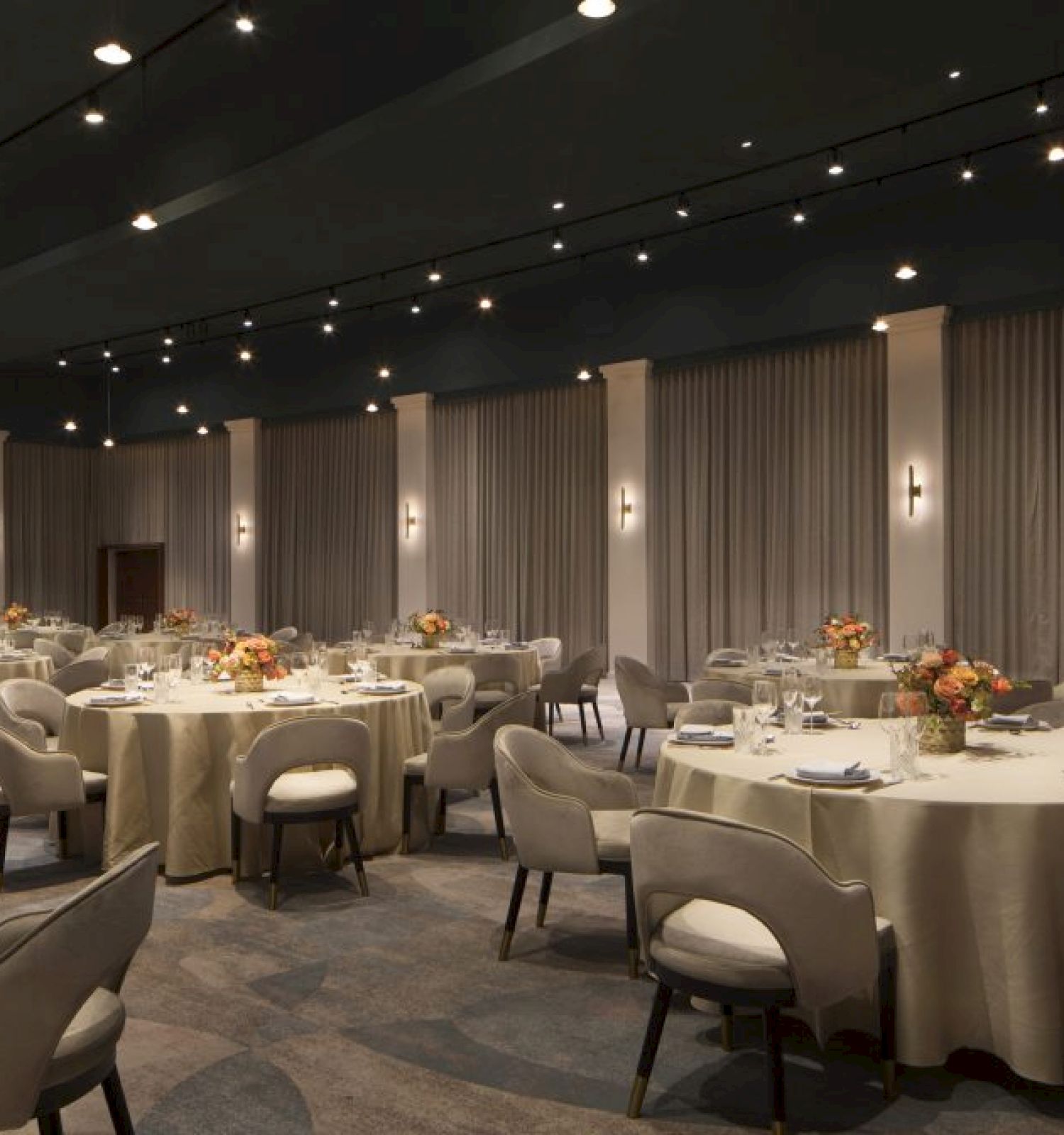 The image shows a banquet hall with round tables covered in beige tablecloths, set with tableware, flowers, and surrounded by cushioned chairs, well-lit environment.