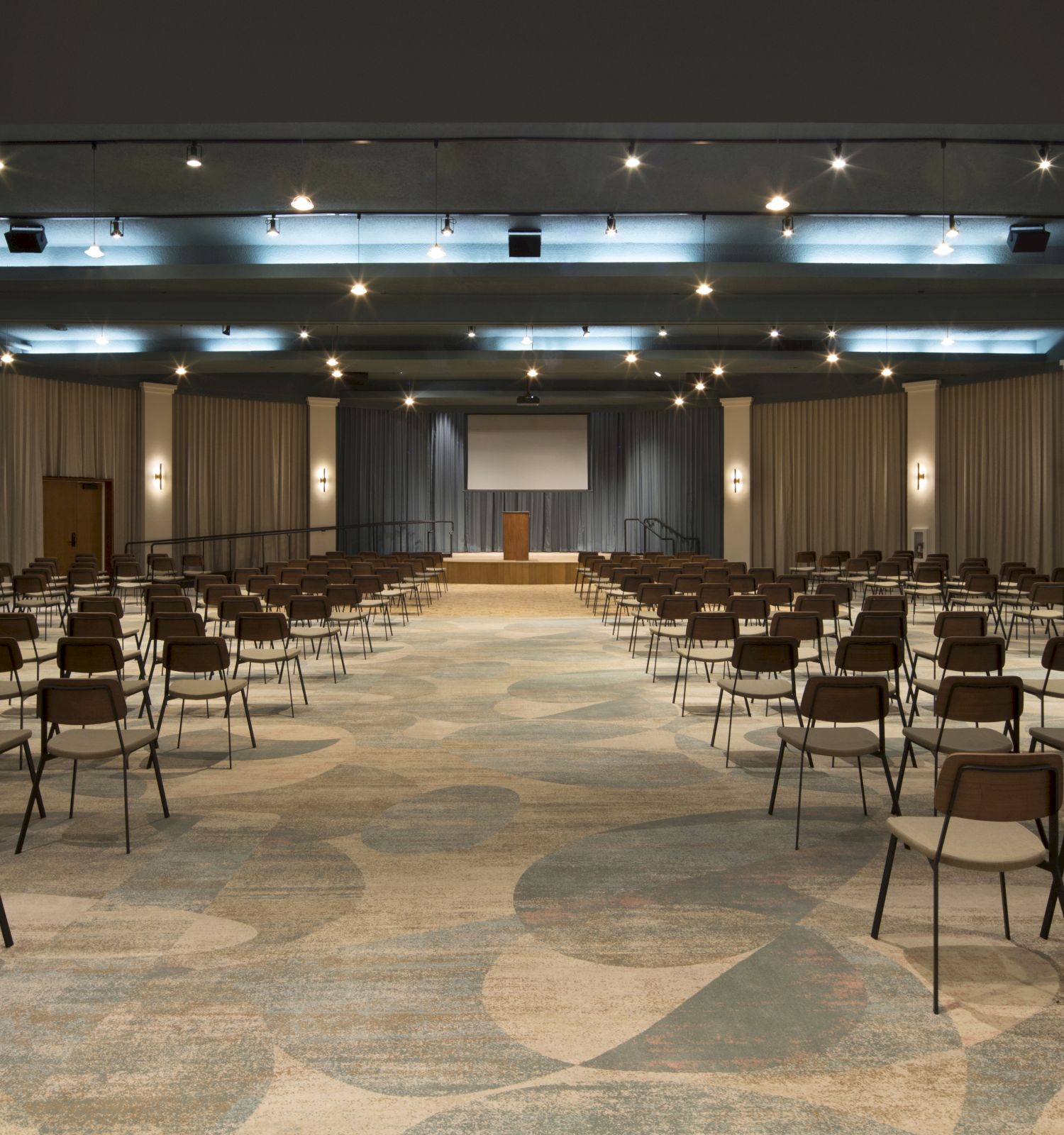 An empty conference hall with rows of chairs, a stage with a podium, and a projection screen at the far end. The lighting is evenly distributed.