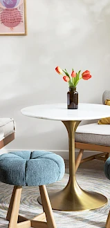 A modern room with a round table, tulips in a vase, blue stools, a bed, armchair, and wall art. The room is bright with natural light.