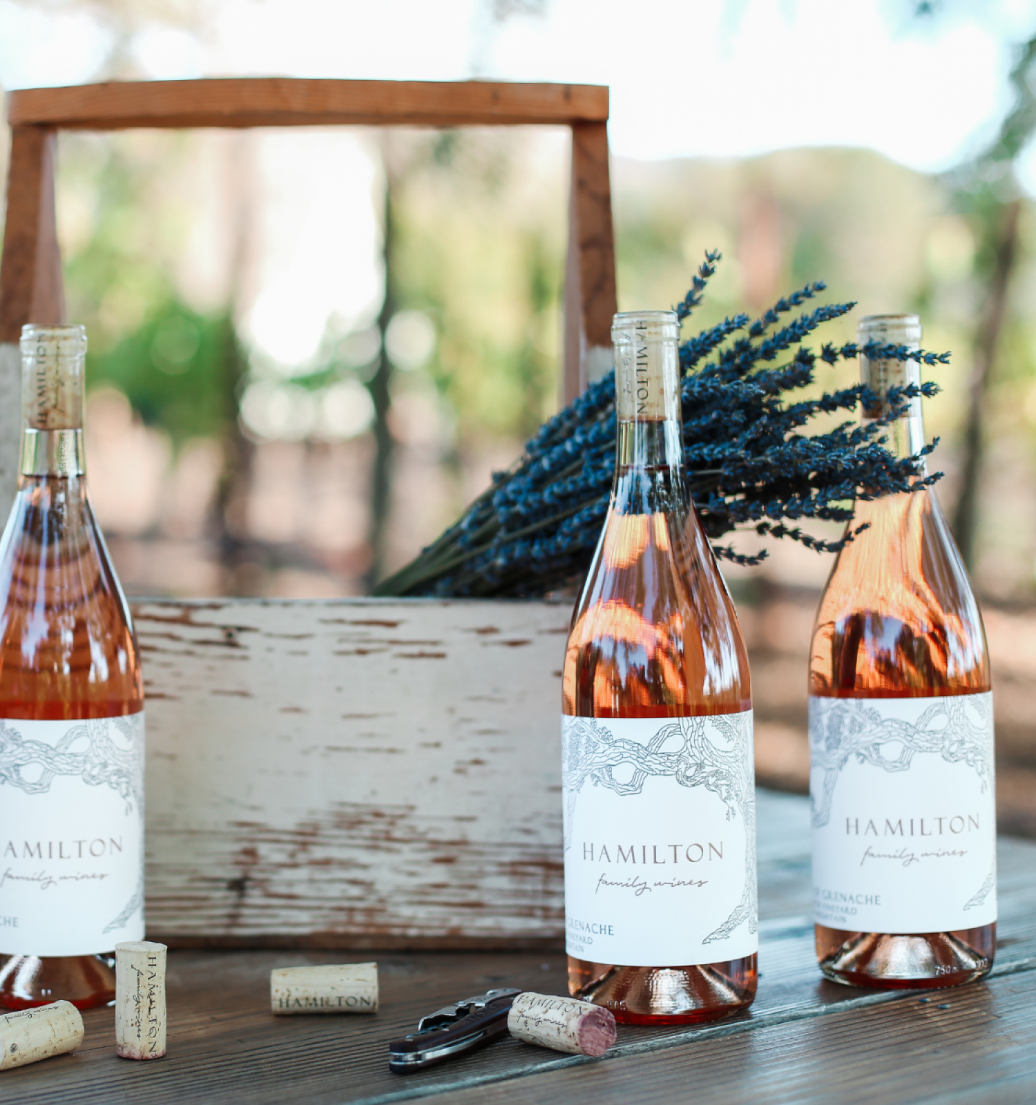 Three bottles of Hamilton wine, some corks, a wine opener, and a rustic wooden box with lavender flowers are arranged on a table.