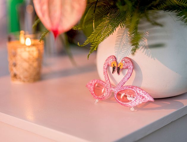 A white vase with pink anthurium flowers on a table, next to a small lit candle and a decorative pink butterfly.