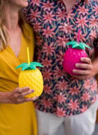 Two people are holding colorful, pineapple-shaped drink cups with straws, wearing bright, summery clothing.