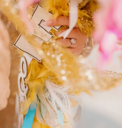 A hand holding colorful, decorative objects, including gold and pink feathers, sequins, and letters. A ring is visible on the hand.