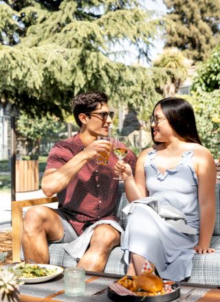 A couple is sitting outdoors, toasting drinks. There's food on the table, and they are enjoying a sunny day together.