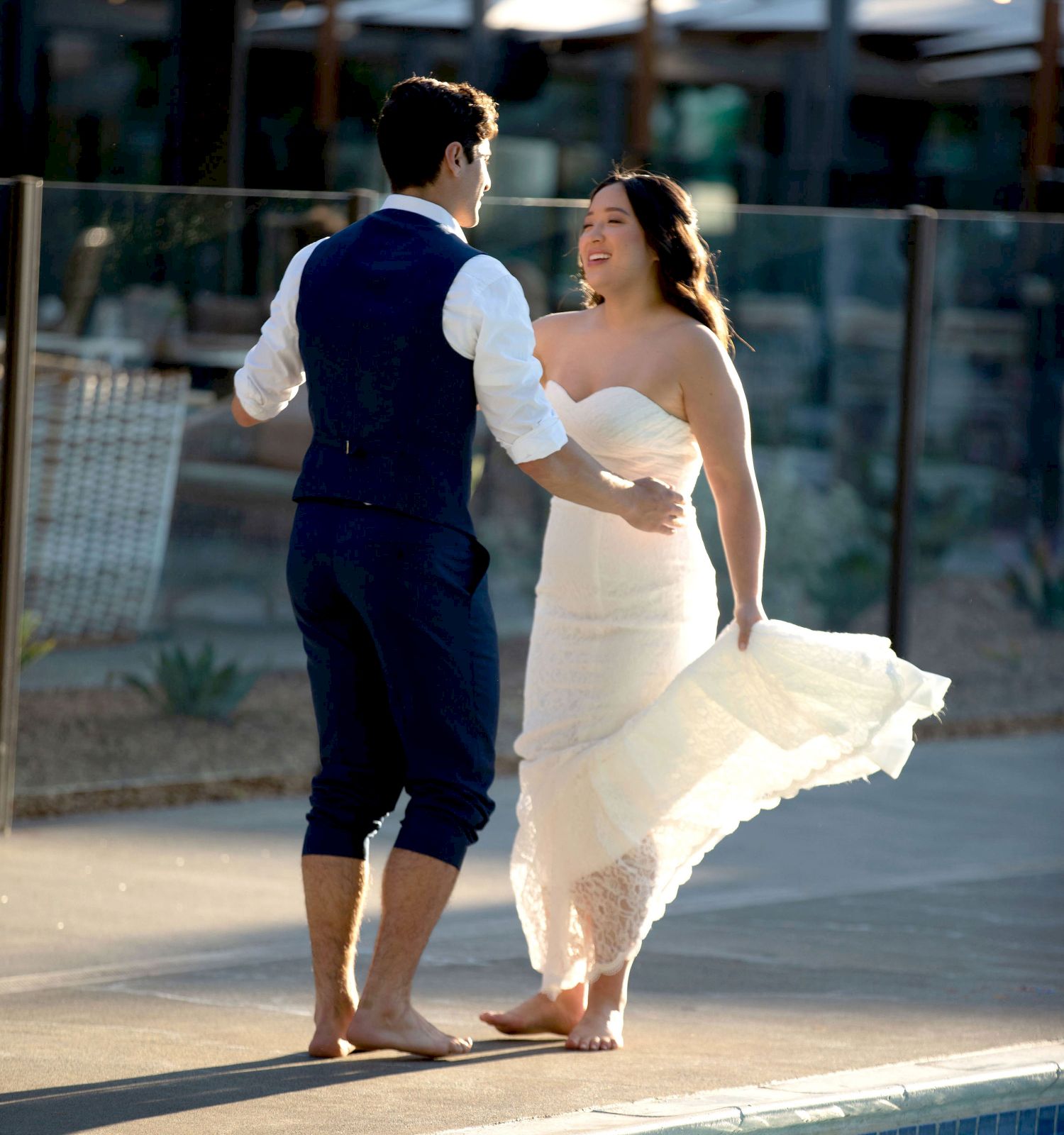 A couple is standing barefoot near a pool, with the man wearing a vest and rolled-up pants and the woman in a white dress, sharing a joyful moment.