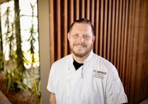 A person in a white chef's coat standing in front of a wooden wall with plants on the side, smiling at the camera.