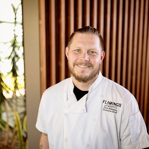 A person in a white chef's coat standing in front of a wooden wall with plants on the side, smiling at the camera.