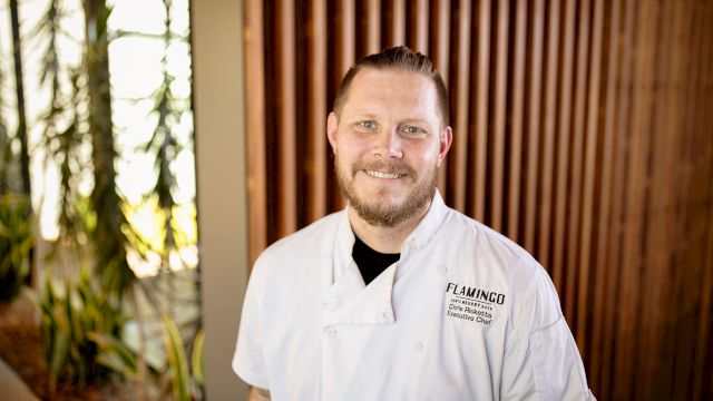 A person in a white chef's coat standing in front of a wooden wall with plants on the side, smiling at the camera.