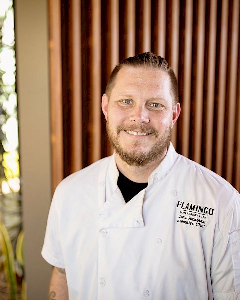 A person in a white chef's coat standing in front of a wooden wall with plants on the side, smiling at the camera.