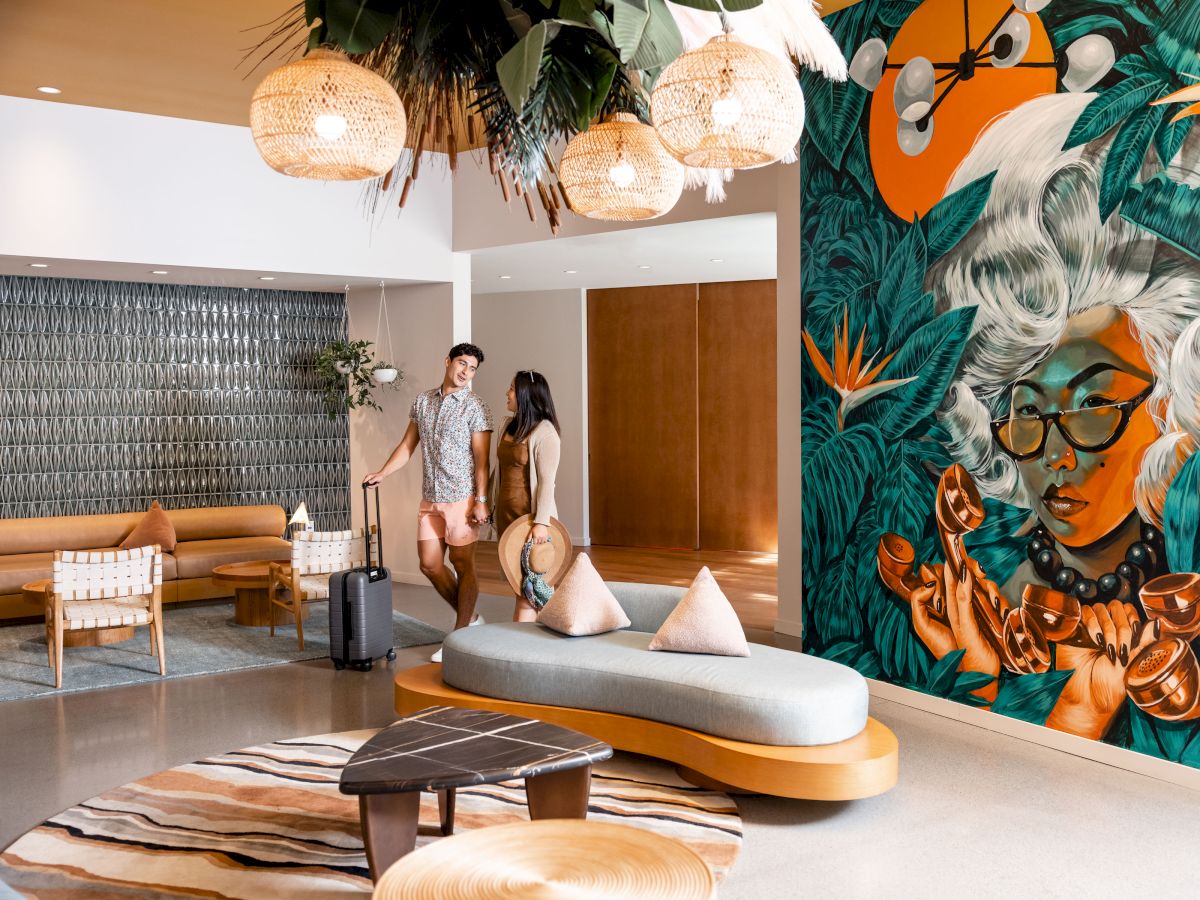 A modern lobby with a large mural of a woman, stylish seating, and two people walking with luggage. Warm, artistic ambiance.