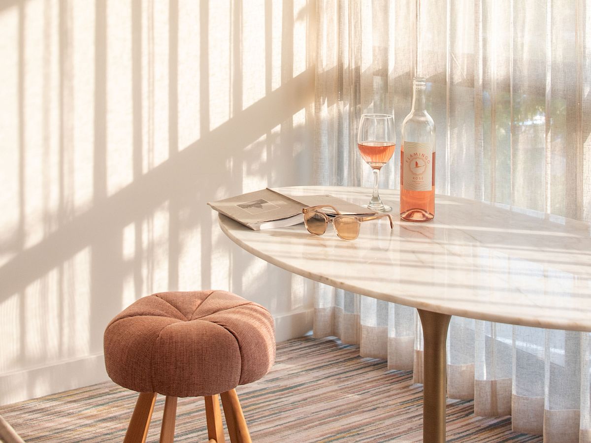 A cozy room with a round table, stool, and open book. Wine and glass on the table, sunlight casting shadows through sheer curtains.