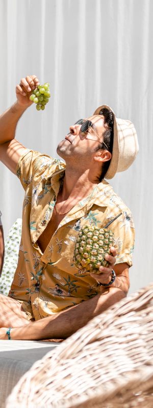 Two people are relaxing on a lounge chair, enjoying tropical fruits and drinks while wearing summer outfits.