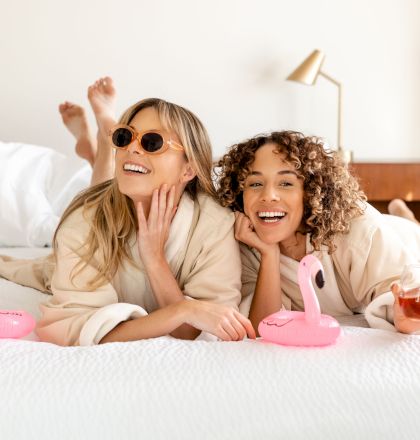 Two women in robes lie on a bed, smiling, with pink flamingo floats and a drink. They look relaxed and are enjoying a pleasant moment together.