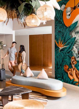 A modern lobby with stylish furniture, a vibrant mural of a woman with white hair and glasses, and two people with luggage near the seating area.