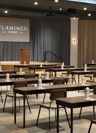 A conference room setup with arranged chairs and desks, featuring bottled water and papers, with a podium and a screen displaying "Flamingo."