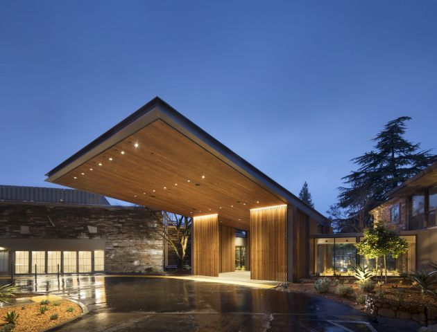 Modern building entrance with wooden canopy, illuminated at night, surrounded by landscaped plants and wet pavement reflecting light.