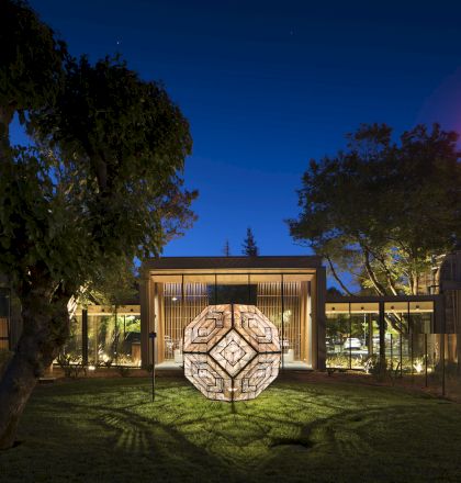 A geometric sculpture illuminated in front of a modern building at night, with trees and a neon hotel sign in the background.