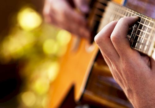 A close-up of hands playing an acoustic guitar, with a blurred natural background and warm lighting illuminating the scene.