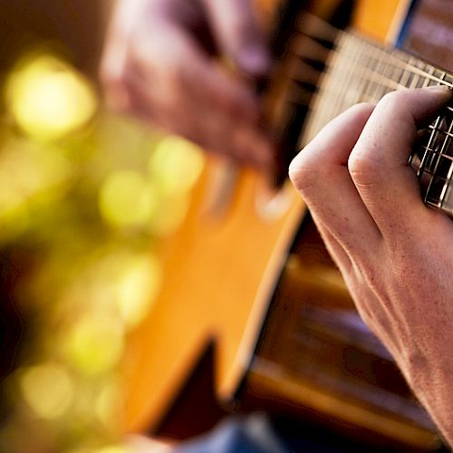 A close-up of hands playing an acoustic guitar, with a blurred natural background and warm lighting illuminating the scene.