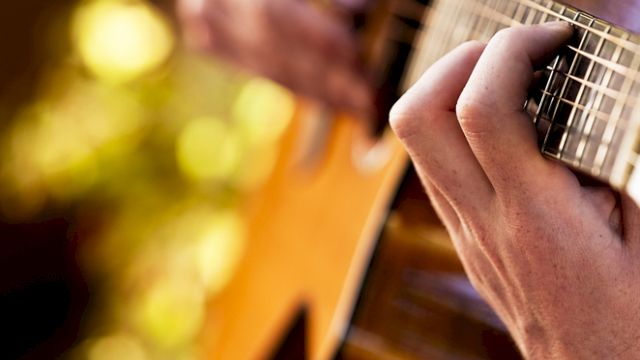 A close-up of hands playing an acoustic guitar, with a blurred natural background and warm lighting illuminating the scene.