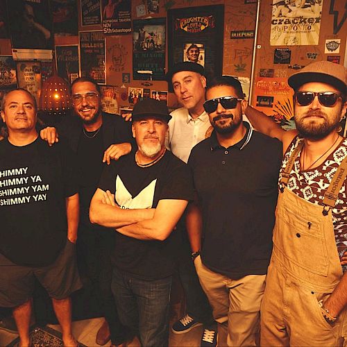 A group of six men pose in a room with posters on the walls. They're casually dressed, and one wears sunglasses.