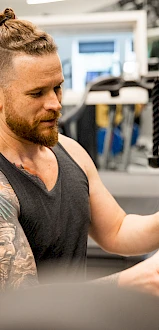 A person with a beard and tattoo is using gym equipment, wearing a tank top. There are treadmills and windows in the background.