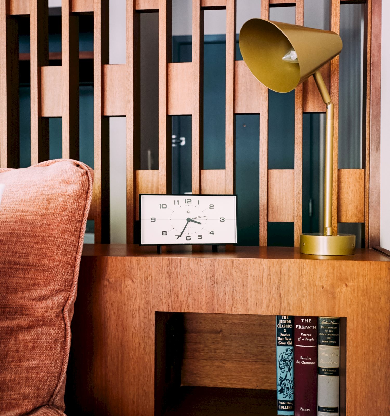A wooden shelf with books, a retro clock, and a gold lamp; a rust-colored pillow nearby adds warmth to the cozy setting.