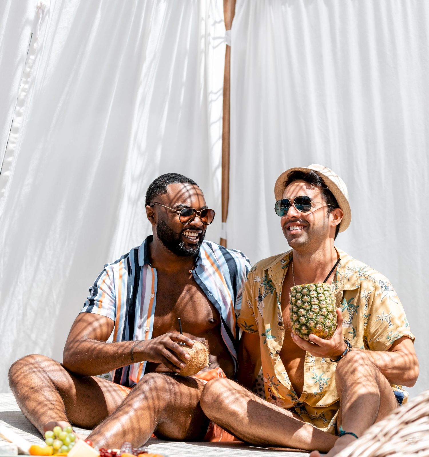 Two people are smiling while sitting together, holding tropical fruits and wearing summer attire under a canopy.
